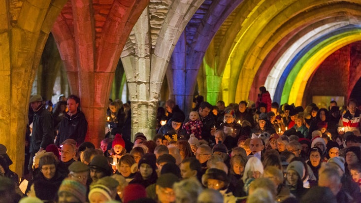 A crowd is gathered to ding Christmas carols among the lit-up ruins of Fountains Abbey, with rainbow lights illuminating the building.